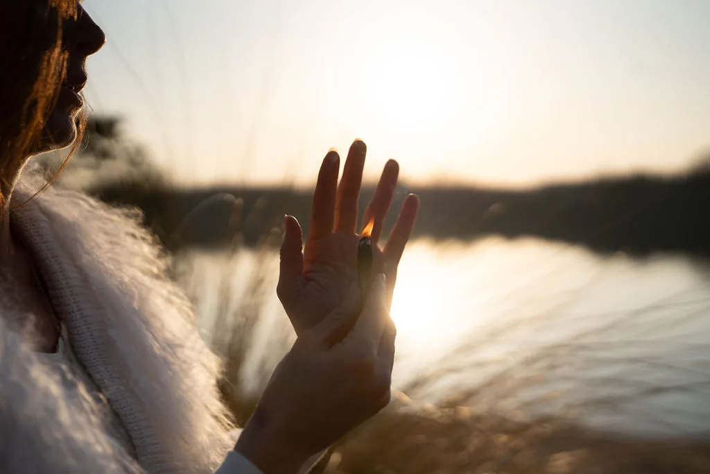 Close-up of Dany Fenton's open hand raised toward a blazing sunset over a lake, golden light streaming through her fingers – a gesture of surrender and breathwork