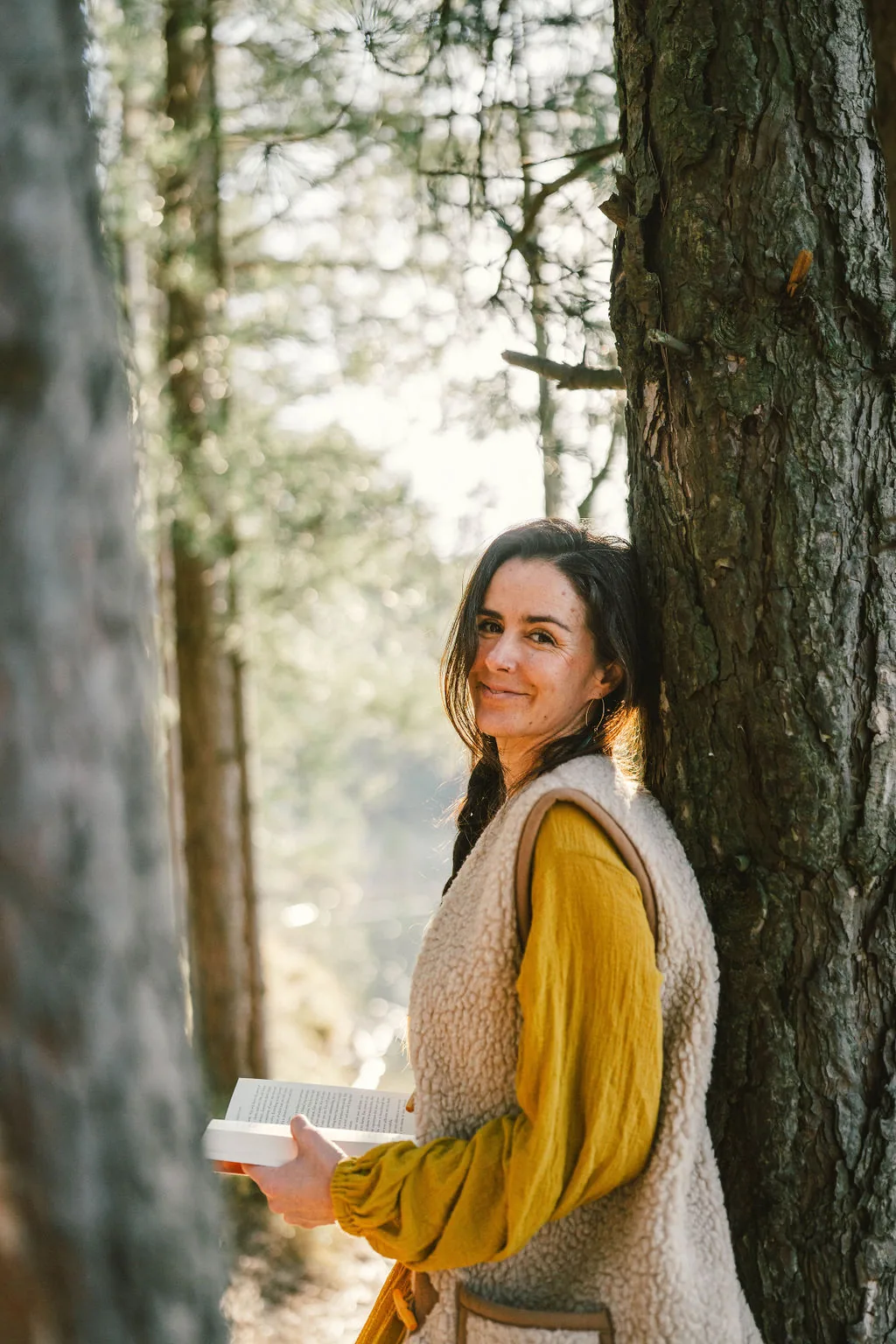 Dany Fenton smiling warmly while leaning against a pine tree in a golden-lit forest, holding a book