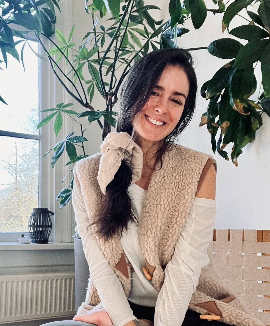 Dany Fenton smiling warmly indoors, seated among large tropical plants in a bright room, wearing a sherpa vest – relaxed, joyful and approachable