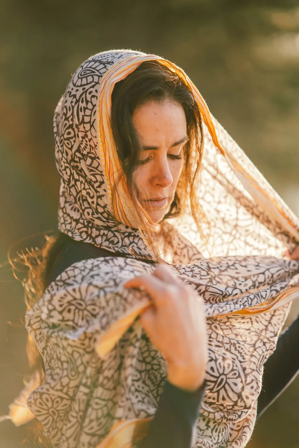 Dany Fenton looking directly at camera, placing a patterned black-and-white shawl over her head in warm golden light – a direct, open invitation to connect