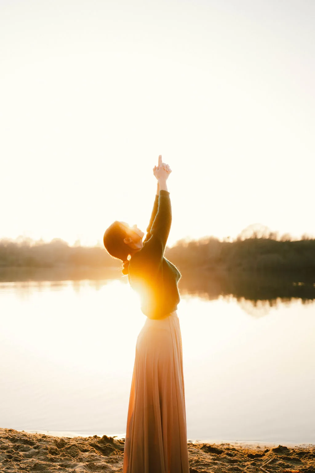 Dany Fenton standing by a lake at sunrise, one arm raised with finger pointing skyward in a Kundalini gesture, skirt flowing – upward energy and possibility