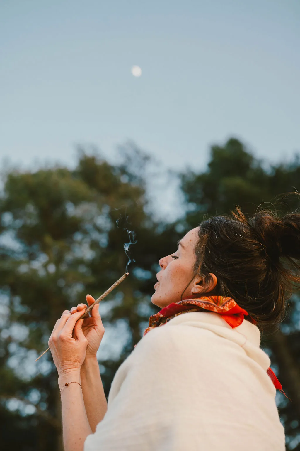 Dany Fenton in profile at dusk, holding a lit incense stick with smoke rising toward a full moon above the pine trees – a ritual moment of breath and intention