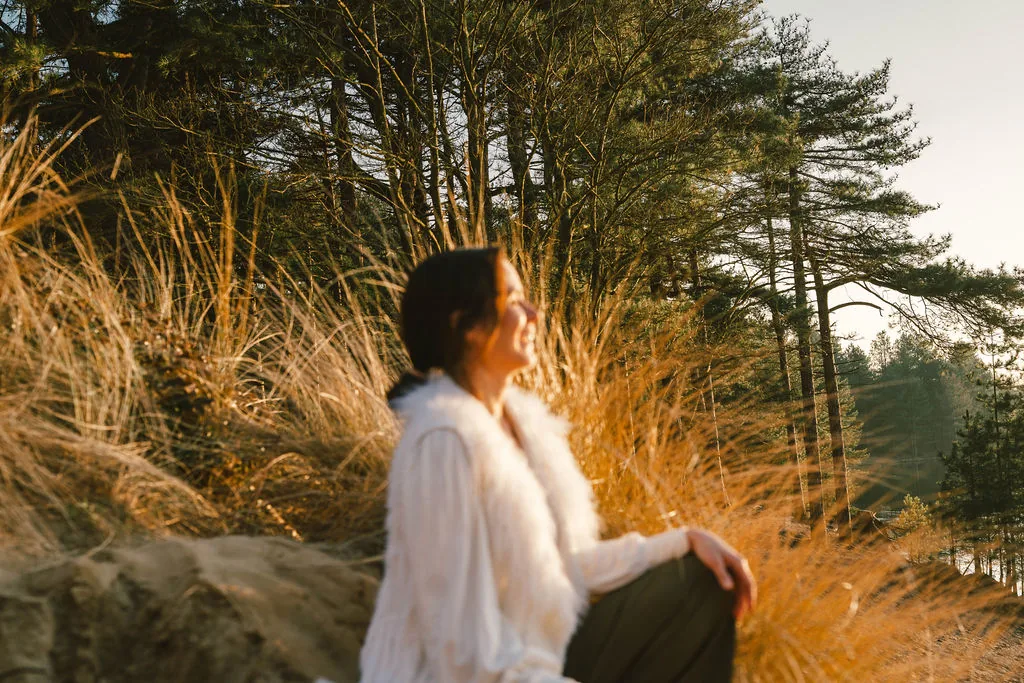 Dany Fenton seated on a sandy dune, smiling and looking upward with golden afternoon light, connecting with the essence of The Reconnected Land