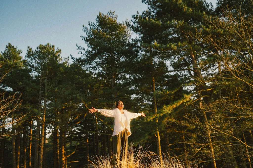 Dany Fenton on a pine-forested hilltop with arms wide open, white fluffy jacket, face tilted skyward
