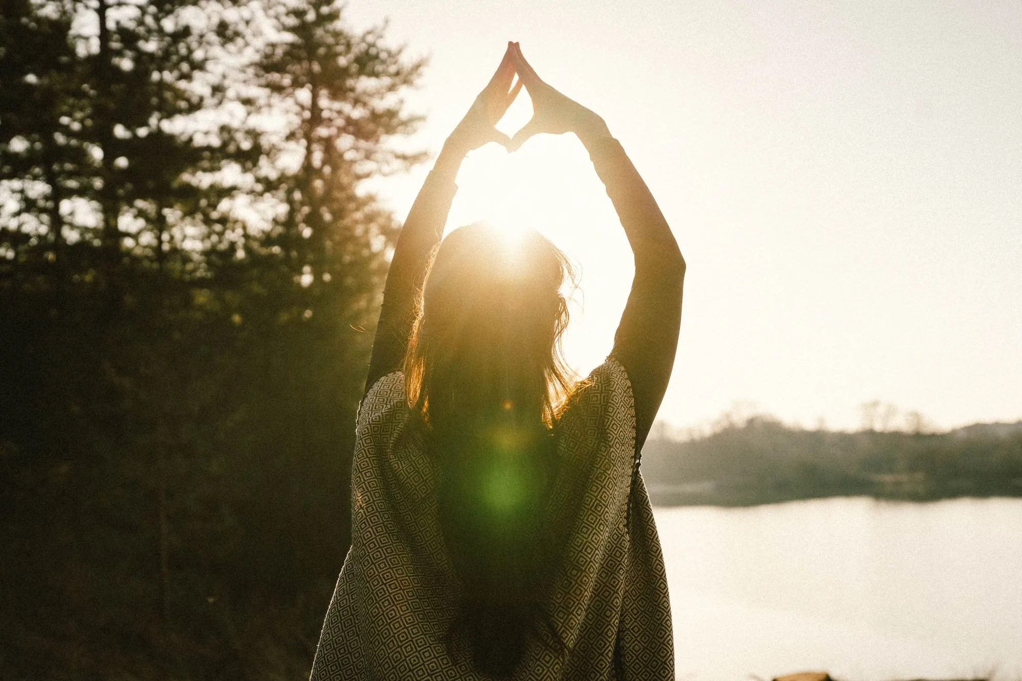 Dany Fenton seen from behind at golden sunset, arms raised overhead forming a heart shape with her hands, patterned shawl, pine trees and lake behind – channelling the Inner Radiance Method