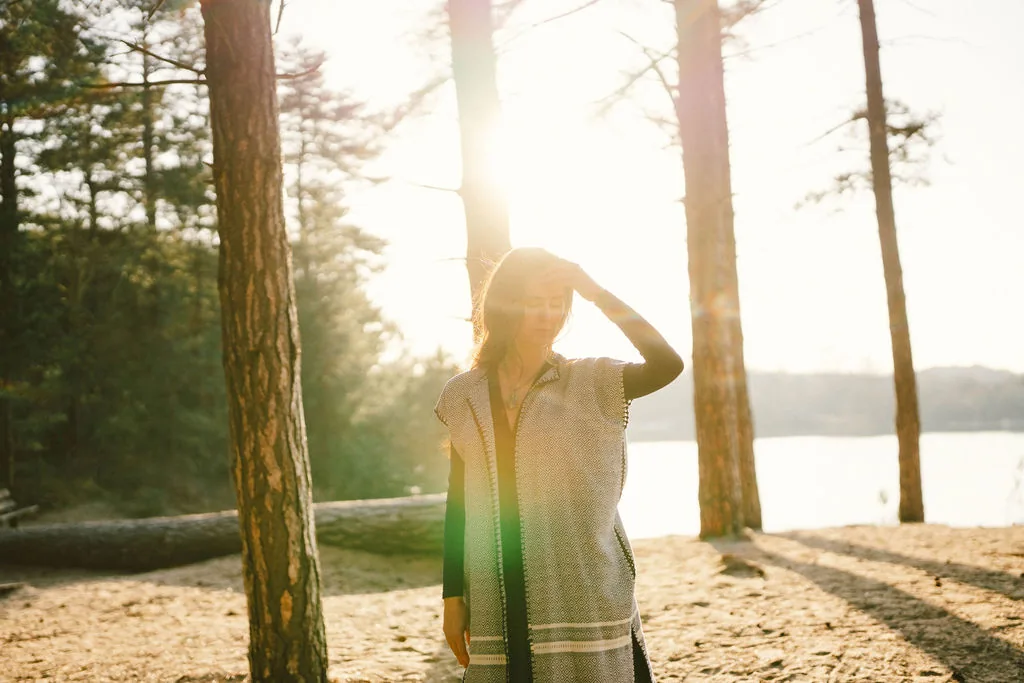 Dany Fenton standing in dappled golden pine forest light, hand raised to her head, patterned shawl, lake visible behind – a moment of internal listening and presence