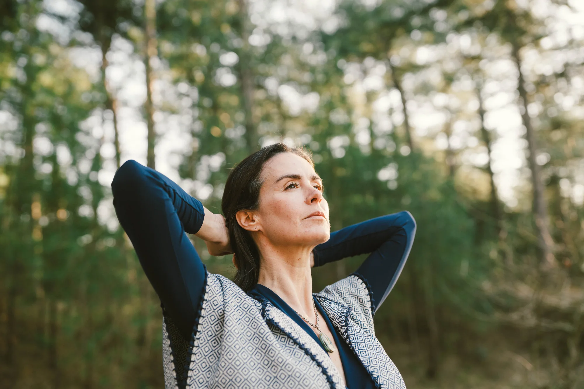 Close portrait of Dany Fenton looking upward in a pine forest, hands clasped behind her head, blue long-sleeve top and diamond-patterned shawl – strong, open and quietly powerful