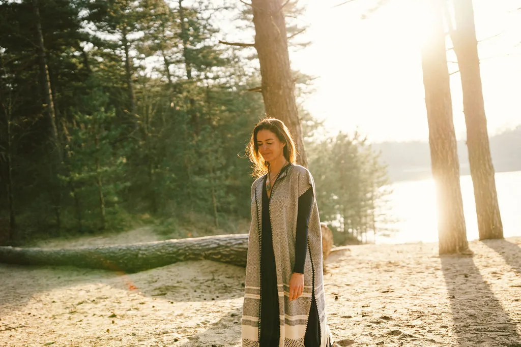 Dany Fenton on a sandy beach at golden sunset, draped in a patterned blanket shawl, walking ahead with quiet intention