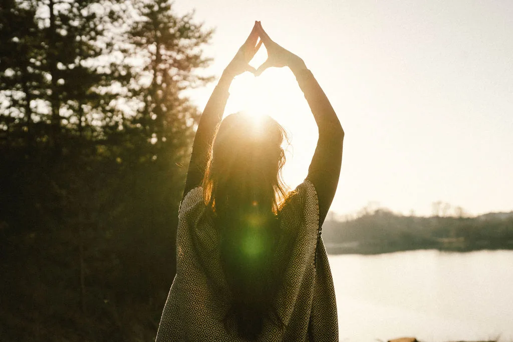 Dany Fenton seen from behind at golden sunset, arms raised overhead forming a heart shape, patterned shawl, lake and pine trees behind – an open-hearted invitation to begin