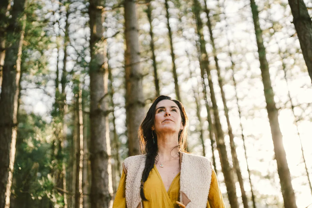 Dany Fenton standing tall among towering pine trees in a golden dress and cream vest, looking upward – embodying the inner strength cultivated in The Reconnected Land's youth programmes