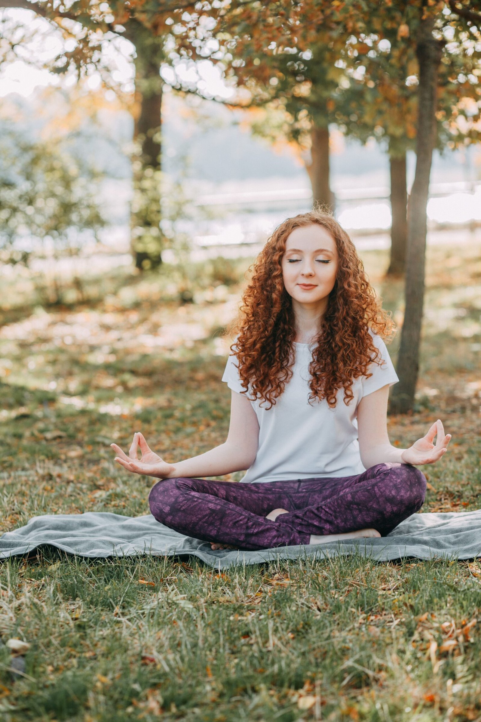 Young teenage girl with curly red hair meditating in lotus pose on a mat on grass, eyes closed peacefully – representing mindfulness practices for teens at The Reconnected Land